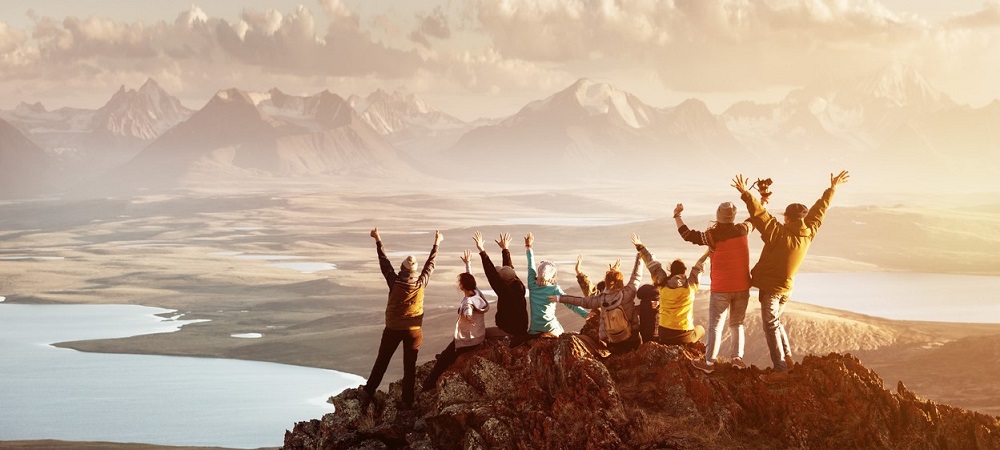 Big group of people having fun in success pose with raised arms on mountain top against sunset lakes and mountains. Travel, adventure or expedition concept